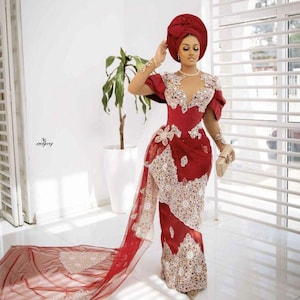 Puede incluir: Un vestido formal rojo con intrincados bordados blancos y dorados. El vestido presenta un escote pronunciado, mangas cortas y una larga cola. Un turbante y un clutch a juego completan el conjunto. Adecuado para ocasiones especiales.