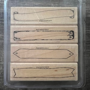 May include: A set of four wooden stamps in a clear plastic case. The stamps feature different banner designs, including a scroll, a plank, and two different ribbon banners.