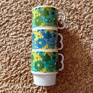 Puede incluir: Tres tazas de cerámica blanca apiladas con un estampado floral en tonos azules, verdes y amarillos. Cada taza tiene un diseño floral único y un asa en forma de corazón. Las tazas están apiladas una encima de la otra.