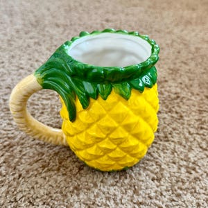 Peut inclure: Une tasse en céramique en forme d'ananas. La tasse est jaune avec une surface texturée, des feuilles vertes en haut et une anse crème. L'intérieur de la tasse est blanc.