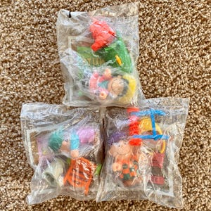 May include: Three sealed plastic bags containing small, colorful toys. The toys appear to be miniature figures with various accessories, including a red helicopter, a green slide, and a small orange chair. The toys are on a brown carpet.