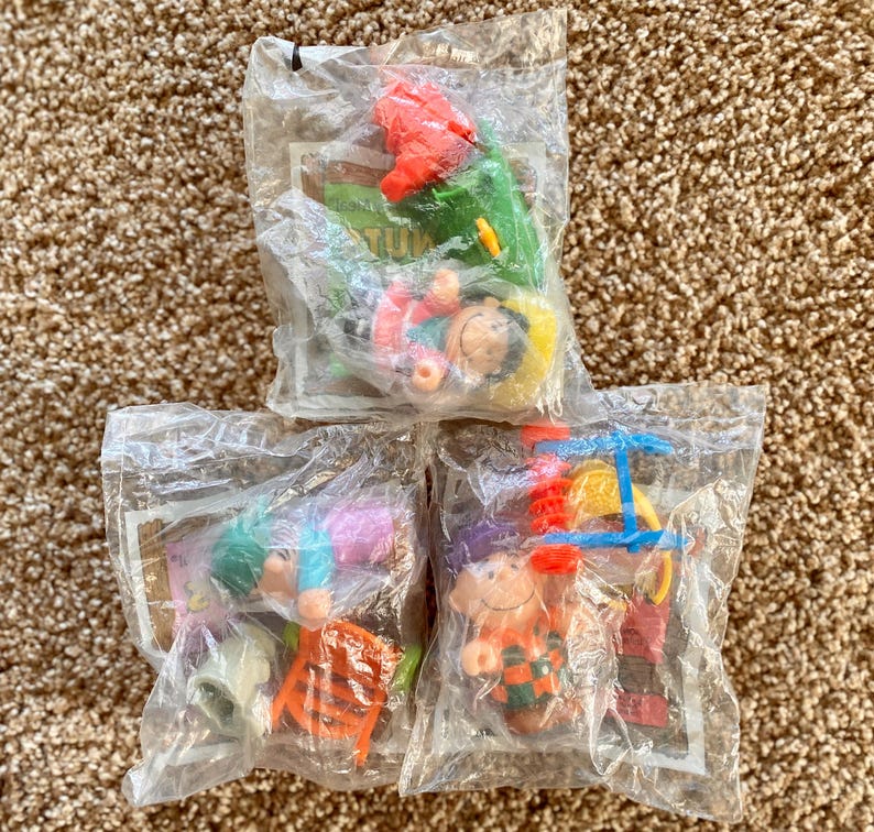 May include: Three sealed plastic bags containing small, colorful toys. The toys appear to be miniature figures with various accessories, including a red helicopter, a green slide, and a small orange chair. The toys are on a brown carpet.