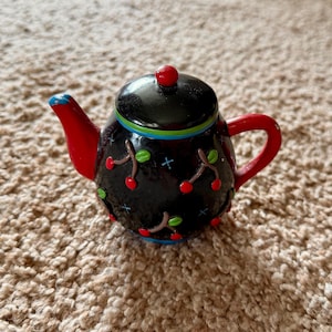 May include: A small, decorative teapot with a black body decorated with red cherries and green leaves. The spout and handle are red, with a blue accent on the spout. The lid is black with a red knob, and the rim has blue and green stripes.