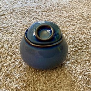 Könnte beinhalten: Ein blaues Keramikgefäß mit Deckel. Das Gefäß hat eine abgerundete Form mit glänzendem Finish und einem dunkelbraunen Rand. Der Deckel hat eine kleinere, abgerundete Form mit einer vertieften Mitte. Das Gefäß steht auf einem beigefarbenen Teppich.