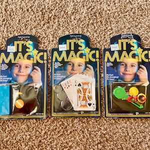 Könnte beinhalten: Drei Vintage-Magier-Sets "It's Magic!" für Kinder ab 6 Jahren. Jedes Set enthält einen anderen Zaubertrick, darunter einen verschwindenden Ball, einen Kartentrick und einen Münzentrick. Die Sets sind in bunten Schachteln mit Illustrationen von Kindern verpackt, die Zaubertricks vorführen.