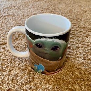 May include: A white ceramic mug featuring a green character with large eyes and a tan robe. The mug has a white handle and a black background. The character is from a popular science fiction franchise.