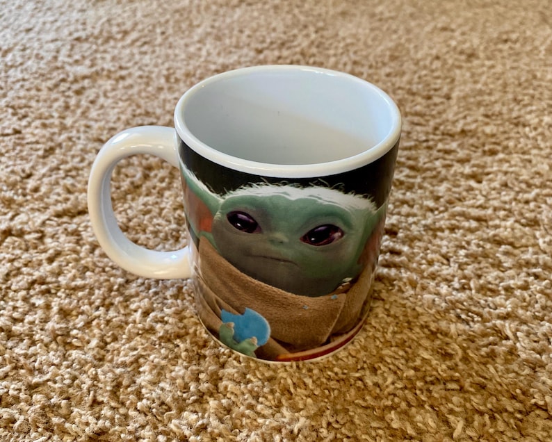 May include: A white ceramic mug featuring a green character with large eyes and a tan robe. The mug has a white handle and a black background. The character is from a popular science fiction franchise.
