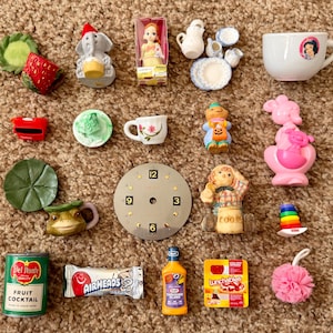 May include: A collection of miniature toys and trinkets, including a doll, teacups, a clock face, a frog mug, and a can of fruit cocktail. Other items include a strawberry, a toy elephant, and a bottle of salad dressing.