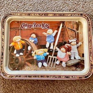 May include: Rectangular metal tray with a Cabbage Patch Kids design. The image on the tray shows several dolls near a wagon wheel and farm tools. The tray has a cream background with a red and gold border.