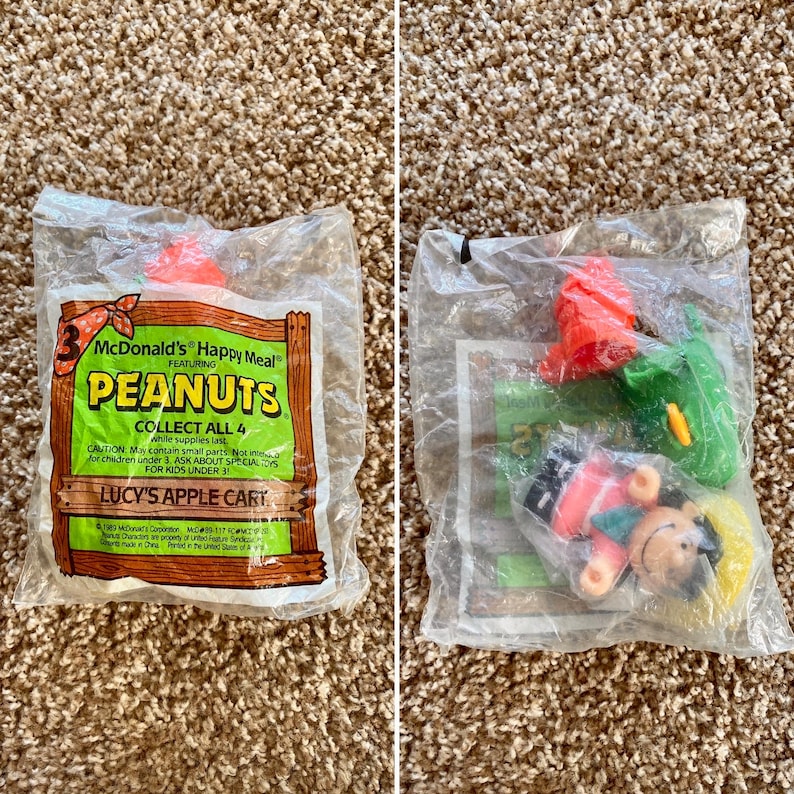 May include: Two sealed McDonald's Happy Meal toys featuring Peanuts characters. One package displays the text "LUCY'S APPLE CART" and "COLLECT ALL 4". The other package contains a Lucy figure, a green item, and a red item.
