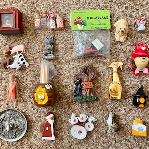 May include: A collection of miniature decorative items, including animal figurines, a red cabinet, a gold spray bottle, and a variety of small bottles. The items are arranged on a textured surface.