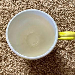 May include: A ceramic coffee mug with a white rim and a yellow handle. The interior of the mug is a pale yellow color. The mug is sitting on a brown, textured surface.