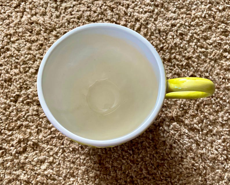 May include: A ceramic coffee mug with a white rim and a yellow handle. The interior of the mug is a pale yellow color. The mug is sitting on a brown, textured surface.