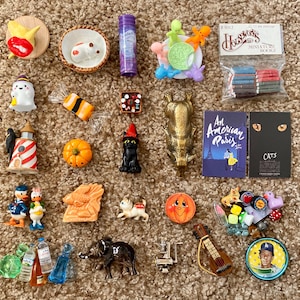 May include: A collection of miniature figurines and trinkets, including a red and yellow lip-shaped pin, a miniature book set, a black cat with a witch hat, and a miniature guitar. Other items include a pumpkin, a ghost, and a miniature book titled "An American Paris."