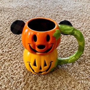 Könnte beinhalten: Eine Halloween-Tasse aus Keramik in Form eines Kürbisses mit einem lächelnden Gesicht. Die Tasse ist orange und gelb mit einem grünen Griff und schwarzen Ohrakzenten. Die Tasse ist so gestaltet, dass sie einer beliebten Zeichentrickfigur ähnelt.