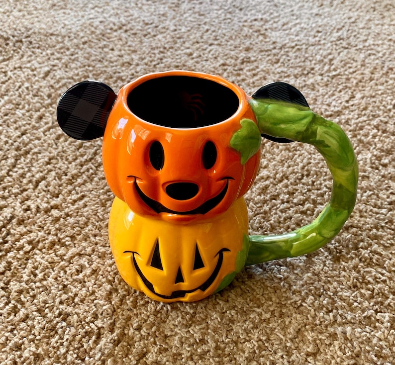 以下が含まれることがあります： ハロウィーンをテーマにしたセラミックマグ。笑顔の顔をしたカボチャの形をしています。マグはオレンジと黄色で、グリーンのハンドルと黒い耳のアクセントが付いています。マグは、人気のある漫画のキャラクターに似せてデザインされています。
