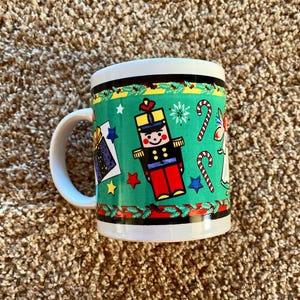 May include: A white ceramic mug with a green, red, and black Christmas design. The design features a nutcracker, candy canes, and stars.