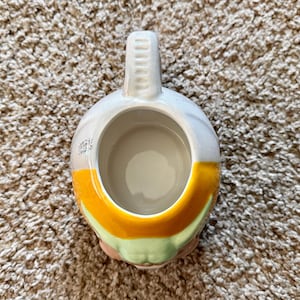 May include: A ceramic mug with a rounded body and a handle. The mug features a design with white, yellow, and green colors. The mug is designed to hold beverages.