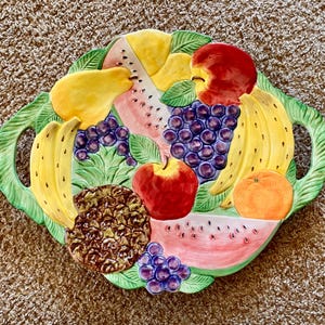 Pode incluir: Uma travessa decorativa de cerâmica com um design de frutas. A travessa apresenta uma variedade de frutas coloridas, incluindo maçãs, bananas, uvas, uma pera, abacaxi, melancia e uma laranja. A travessa tem uma borda verde em forma de folha com alças.