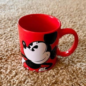 K&ouml;nnte beinhalten: Eine rote Keramik-Tasse mit einem Mickey Mouse-Design. Die Tasse hat einen gro&szlig;en Henkel und ist f&uuml;r hei&szlig;e oder kalte Getr&auml;nke geeignet. Die Disney-Figur ist in Schwarz, Wei&szlig; und Rot dargestellt.