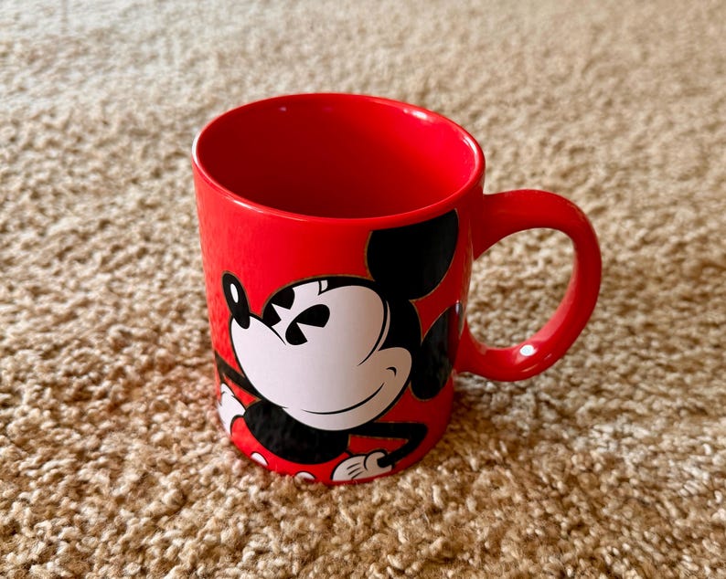 K&ouml;nnte beinhalten: Eine rote Keramik-Tasse mit einem Mickey Mouse-Design. Die Tasse hat einen gro&szlig;en Henkel und ist f&uuml;r hei&szlig;e oder kalte Getr&auml;nke geeignet. Die Disney-Figur ist in Schwarz, Wei&szlig; und Rot dargestellt.