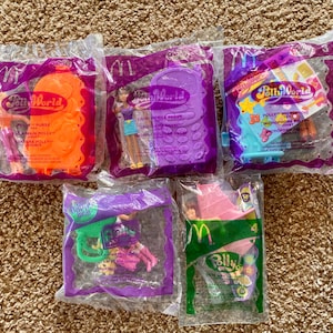 May include: Five sealed McDonald's Happy Meal toys featuring Polly Pocket characters. The toys are in clear plastic bags with purple and green accents. The toys include a purse, a mobile phone, a microphone, and other accessories.