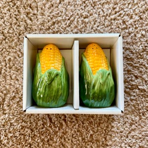 May include: A pair of ceramic salt and pepper shakers shaped like ears of corn, featuring bright yellow kernels and textured green husks. The two shakers are nestled side by side within a white cardboard box with a central divider.