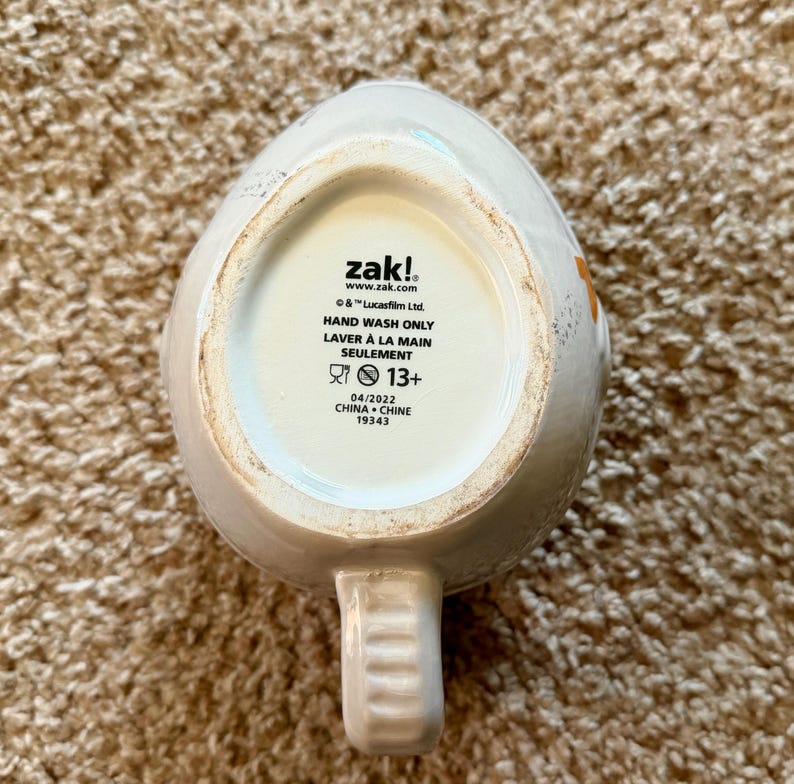 May include: A white ceramic mug with a handle, featuring the brand name "zak!" and the text "HAND WASH ONLY". The bottom of the mug also includes the text "&copy; & &trade; Lucasfilm Ltd." and "04/2022 CHINA CHINE 19343".