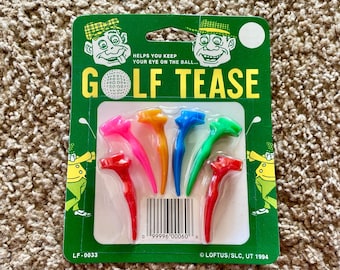 Può includere: Un pacchetto di sei tee da golf in plastica colorati con il testo "GOLF TEASE" e "HELPS YOU KEEP YOUR EYE ON THE BALL..." sul davanti. Il pacchetto include anche un codice a barre e il testo "LOFTUS/SLC, UT 1994".