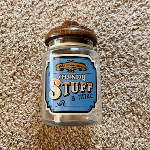 May include: A clear glass storage jar with a dark brown wooden lid. The front features a blue label with the text Olde Thompson est. 1887, Handy Stuff & Misc. in white and orange lettering. The jar has a cylindrical shape and a smooth glass finish.