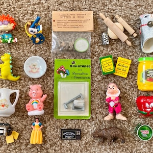 Könnte beinhalten: Eine Sammlung von Miniaturfiguren und Ornamenten, darunter ein gelber Dinosaurier, ein rosa Care Bear, eine Schneewittchen-Figur und ein Miniatur-Apfelmusglas. Weitere Artikel sind Nudelrollen, eine Katzenfigur und kleine Bücher.