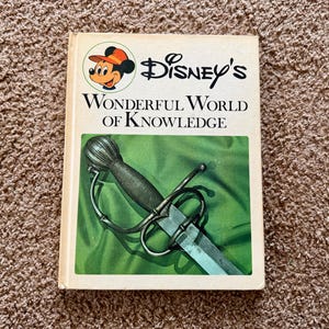 May include: A hardback book titled Disney's Wonderful World of Knowledge with a white cover featuring a circular illustration of Mickey Mouse and a central green rectangular image of a metallic sword with a detailed hilt and crossguard.