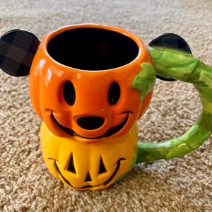 以下が含まれることがあります： 笑顔の顔をしたカボチャの形をしたセラミックハロウィーンマグ。マグはオレンジと黄色で、黒のアクセントと緑のハンドルが付いています。耳は黒で、チェック柄です。秋の季節にぴったりの、楽しくてお祝いのアイテムです。