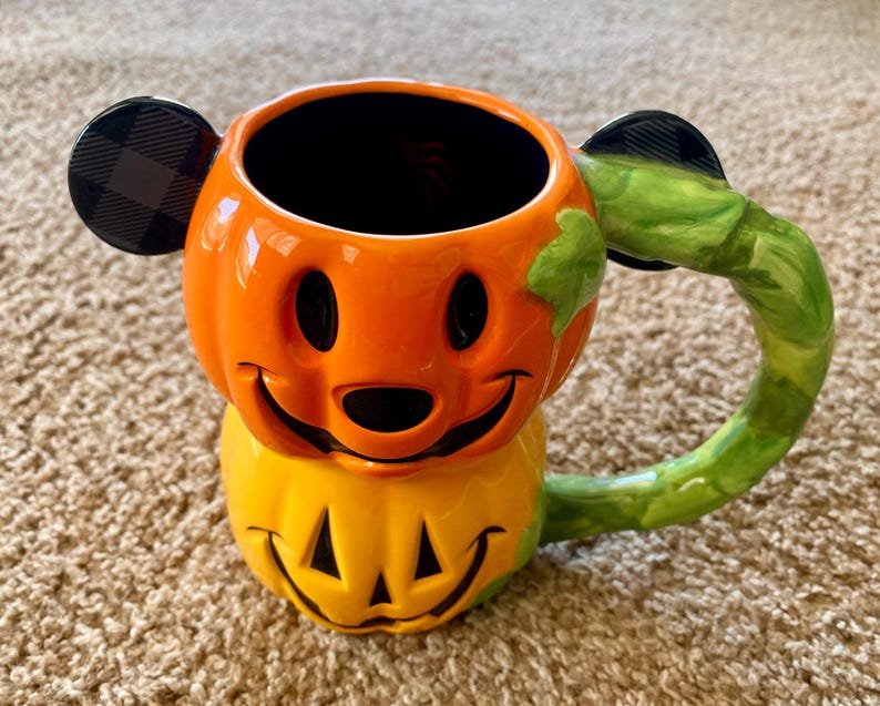 以下が含まれることがあります： 笑顔の顔をしたカボチャの形をしたセラミックハロウィーンマグ。マグはオレンジと黄色で、黒のアクセントと緑のハンドルが付いています。耳は黒で、チェック柄です。秋の季節にぴったりの、楽しくてお祝いのアイテムです。