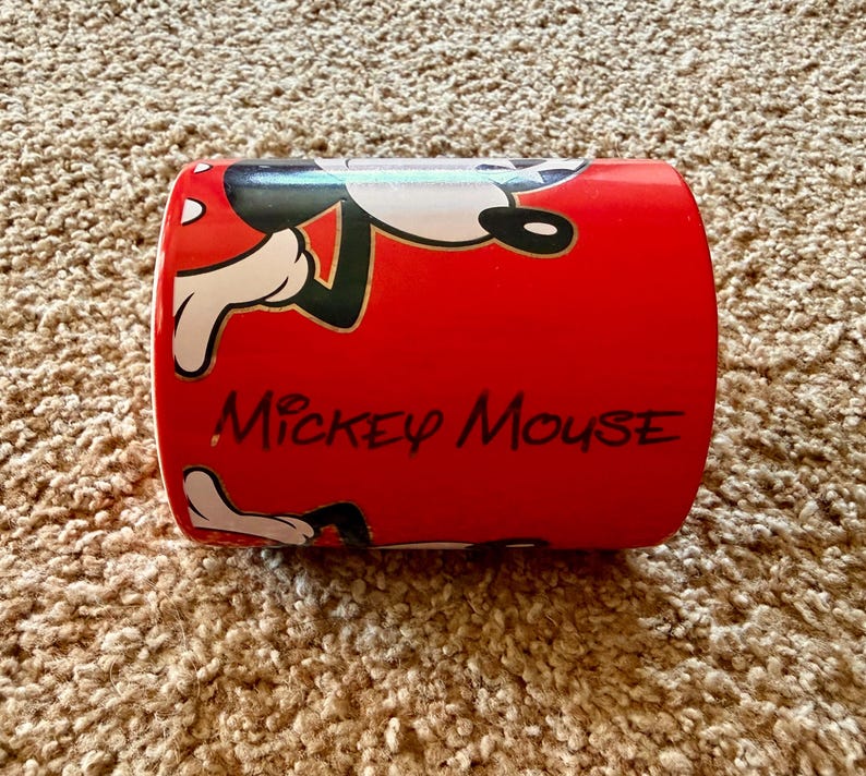 K&ouml;nnte beinhalten: Eine wei&szlig;e Keramik-Tasse mit schwarzem Henkel und dem Text "But First, Coffee" in fetter, schwarzer Schrift. Die Tasse steht vor einem verschwommenen Hintergrund, der eine gem&uuml;tliche Umgebung suggeriert.