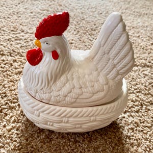 May include: White ceramic chicken figurine with a red comb and wattles, sitting in a white ceramic nest.