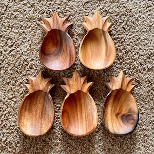 May include: Five wooden pineapple-shaped bowls with a natural wood grain finish. The bowls have a carved design, with a textured top resembling a pineapple crown. The bowls are arranged on a brown, textured surface.