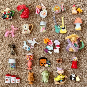 May include: A collection of miniature figurines and charms, including a strawberry pitcher, popcorn, guitars, a clock, a lamp, a gumball machine, and a mushroom. The items are in various colors and styles, arranged on a brown surface.