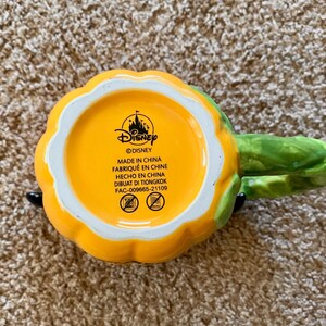 以下が含まれることがあります： カボチャの形をしたセラミックマグ。緑色のハンドルと黄色とオレンジ色の外観。マグカップの底は黄色で、ディズニーのロゴと「MADE IN CHINA」の文字が描かれています。