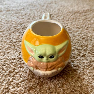 May include: A ceramic mug featuring a character from a popular science fiction series. The mug has a rounded shape with an orange exterior and a light green character face. The character has large ears and black eyes. The handle is white.