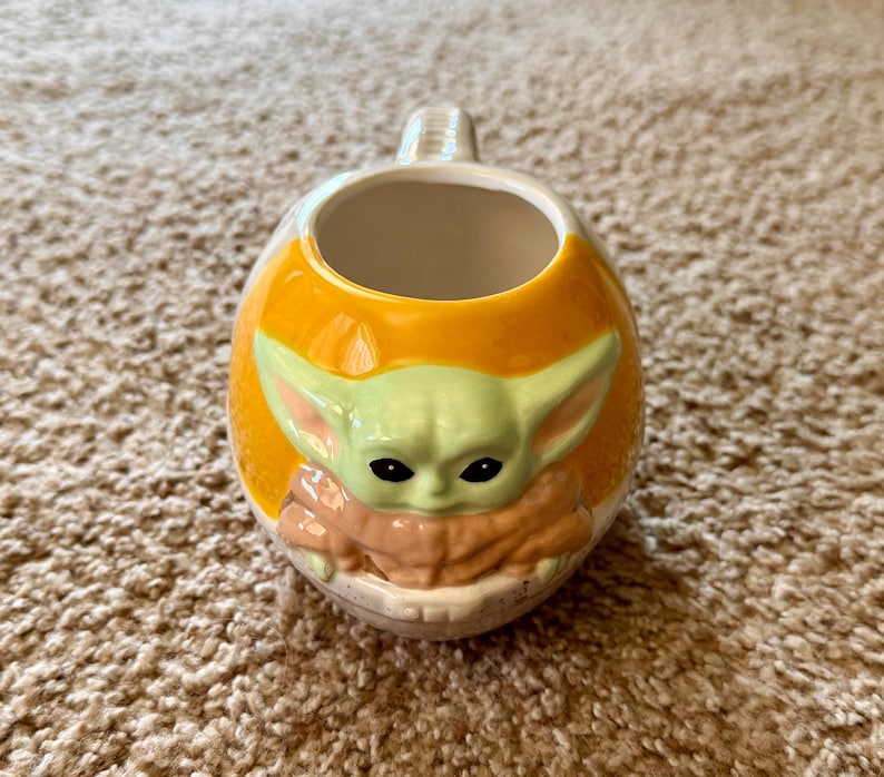 May include: A ceramic mug featuring a character from a popular science fiction series. The mug has a rounded shape with an orange exterior and a light green character face. The character has large ears and black eyes. The handle is white.