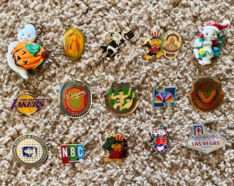 Op de afbeelding: Een verzameling kleurrijke emaille pins met diverse ontwerpen. Inclusief een pompoen-thema spook, een religieus icoon, een panda in smoking, sportteam logo's en een "Welcome to Las Vegas" bord. Deze pins zijn waarschijnlijk verzamelobjecten.