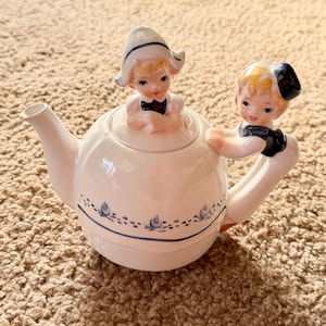 Puede incluir: Una tetera de cerámica blanca con una tapa con una figura de un niño con un sombrero holandés. Otra figura está en el asa. La tetera tiene detalles florales azules y un pico. La tetera está sobre una superficie marrón.