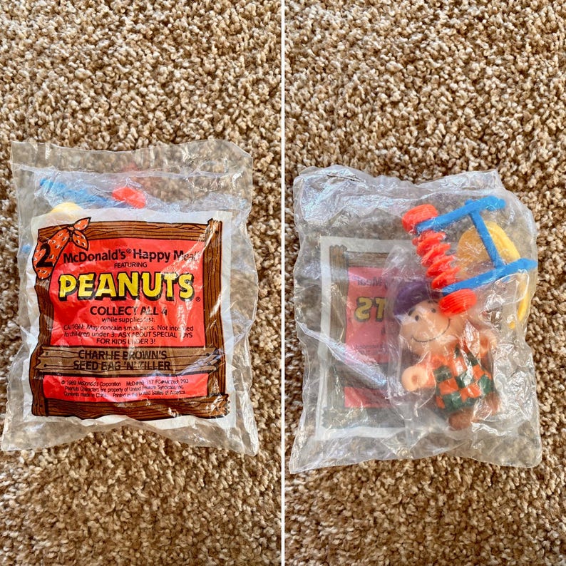 May include: Two sealed McDonald's Happy Meal toys featuring Peanuts characters. One package has a red and brown design with text. The other package contains a toy with a figure wearing a purple hat and orange and green clothing.