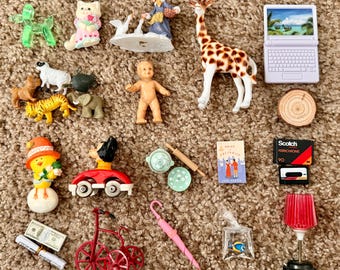 May include: A collection of miniature toys, including a giraffe, a laptop, a baby doll, a red tricycle, and a miniature book. The toys are arranged on a brown, textured surface.