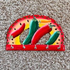 May include: A decorative key holder featuring a vibrant design of chili peppers. The key holder has a red base with four silver hooks and a colorful arch with red, yellow, and green peppers. The peppers are red and green.