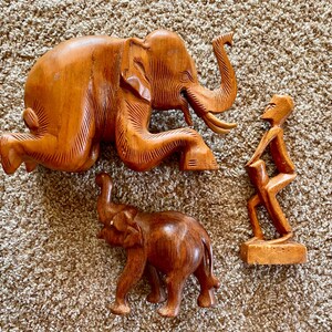 May include: Three wooden sculptures of elephants and a person. The larger elephant is lying down with its trunk raised. The smaller elephant is standing with its trunk down. The person is standing with their arms raised.