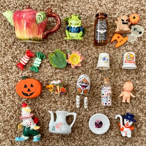 Op de afbeelding: Een verzameling miniatuur decoratieve items, waaronder een theepot, kikkerbeeldje, snoep, pompoen en een fles met de tekst "Wool of Magic". Andere items zijn een vingerhoed, een sneeuwpop en een babyfiguur.