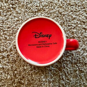 K&ouml;nnte beinhalten: Rote Keramik-Tasse mit wei&szlig;em Rand und rotem Henkel. Der Boden der Tasse zeigt das Disney-Logo und den Text "Microwave and Dishwasher Safe Made in China".