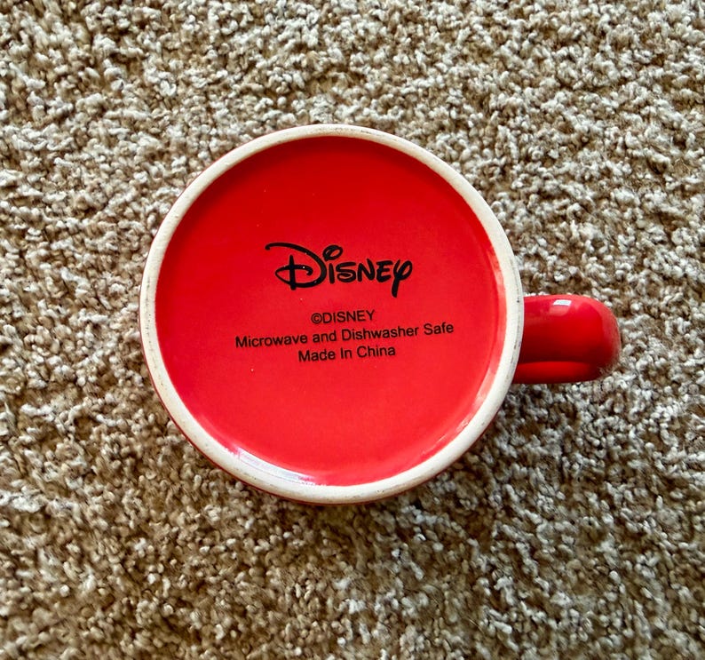 K&ouml;nnte beinhalten: Rote Keramik-Tasse mit wei&szlig;em Rand und rotem Henkel. Der Boden der Tasse zeigt das Disney-Logo und den Text "Microwave and Dishwasher Safe Made in China".